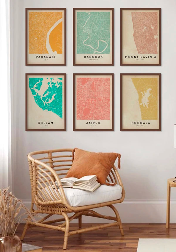 Cozy reading corner with a rattan armchair and a gallery wall of colorful Wanderer Maps™ by Cha, featuring minimalist city maps of Varanasi, Bangkok, Mount Lavinia, Kollam, Jaipur, and Koggala in soft vintage tones.