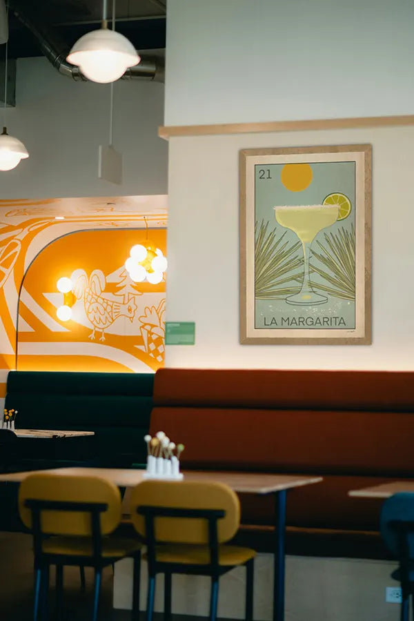 Stylish restaurant interior with mid-century seating and vibrant wall art, featuring the Spanish Capsule™ “La Margarita” poster by Cha, a retro cocktail print inspired by Mexican Lotería cards, with a minimalist glass and agave illustration.