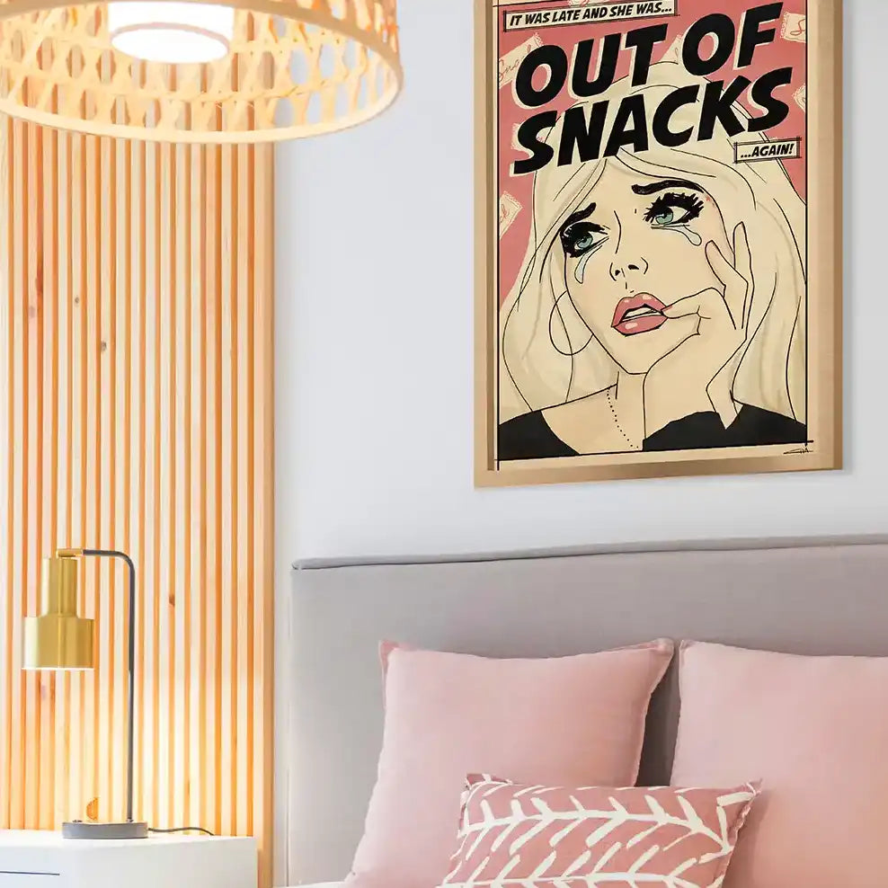 Framed comic-style art print “Out of Snacks” hanging above a pastel pink and white bed in a cozy modern bedroom
