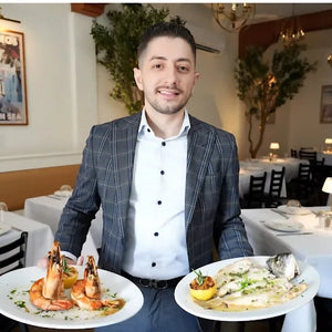 Restaurant owner Granit Shala at Kyria in Harrison NY presenting two Greek signature dishes, with Alecse’s “Petros Mykonos” vintage travel poster from MyRetroposter in the background.