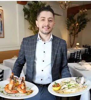 Restaurant owner Granit Shala at Kyria in Harrison NY presenting two Greek signature dishes, with Alecse’s “Petros Mykonos” vintage travel poster from MyRetroposter in the background.