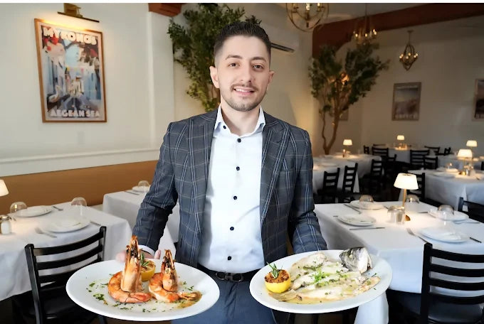 Restaurant owner Granit Shala at Kyria in Harrison NY presenting two Greek signature dishes, with Alecse’s “Petros Mykonos” vintage travel poster from MyRetroposter in the background.