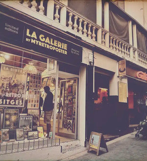 View of La Galerie by Myretroposter in Sitges with people lining up for waffles at the Gofras stand next door – the real-life inspiration behind the Sitges Waffles Poster by Cha. Limited edition food art print.