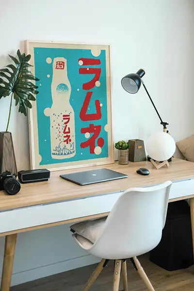 Limited edition ‘Ramune’ Japanese poster by Cha from the Vintage Exotics™ Collection, featuring a retro-style illustration of a classic Ramune soda bottle with bold red Japanese characters against a vibrant blue background, displayed on a modern wooden desk setup with a lamp, plants, and a laptop