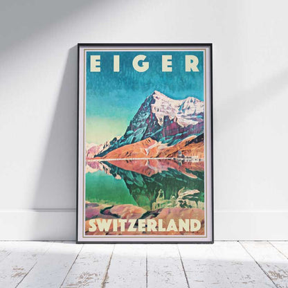 Eiger mountain travel poster in black frame on a wooden floor, capturing a colorful scene of the Swiss Alps with mirror lake reflection