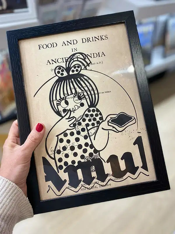 A hand holding a framed vintage-inspired poster featuring the iconic Amul girl, a beloved mascot from Indian advertising. Part of The Great Indian Décor™ collection by Cha, this artwork blends nostalgic typography with retro aesthetics, incorporating elements from an old book cover titled Food and Drinks in Ancient India. The distressed background and monochrome illustration enhance its vintage appeal, making it a unique decor piece.