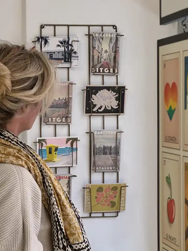 A woman browsing a stylish metal display of postcard packs at La Galerie by Myretroposter in Sitges. The collection features vintage-inspired designs celebrating iconic destinations like Sitges and Barcelona, as well as botanical and pop-art motifs. The carefully curated selection captures the charm of retro travel posters in a smaller format, perfect for collectors or as unique souvenirs.