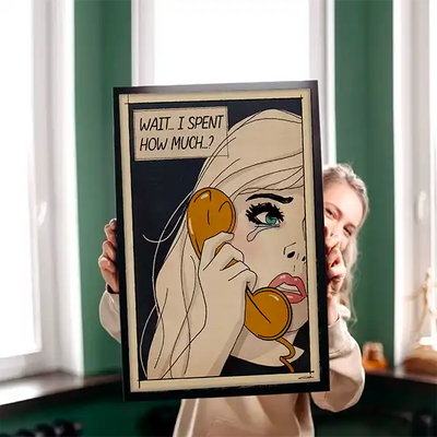 Young woman holding a framed Soft Pop Studio™ art print by Cha, featuring a retro comic-style blonde character saying ‘Wait… I spent how much?’ with bold lines and vintage tones.