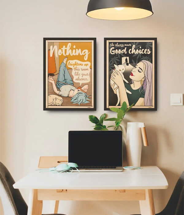 Two framed posters from the Soft Pop Studio™ collection by Cha, featuring retro-inspired illustrations: one with a woman lying with a cat and the text “Nothing brightens up this room like your absence,” and the other depicting a woman holding a cat with the caption “She always made good choices,” displayed above a minimalist desk setup