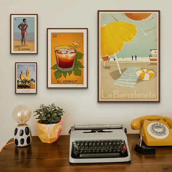 Retro home office setup featuring framed Spanish Capsule™ art prints by Shree (Cha), including playful vintage posters of La Barceloneta beach, a glass of vermut, Sitges skyline, and El Catrín. Displayed above a wooden desk with a yellow rotary phone, vintage typewriter, and ceramic lamp.