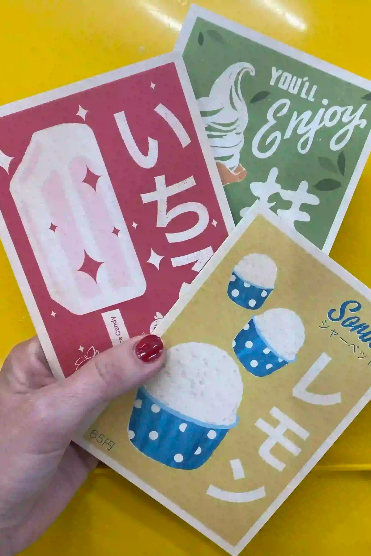 A hand holding three Vintage Exotics™ postcards by Cha, inspired by Japanese retro packaging design. The illustrations feature nostalgic sweet treats: a pink strawberry ice candy, a yellow lemon sorbet in blue polka-dot cups, and a green matcha soft-serve with playful typography and Japanese characters. The textured paper and muted colors evoke a vintage aesthetic, enhancing the collection’s charm.