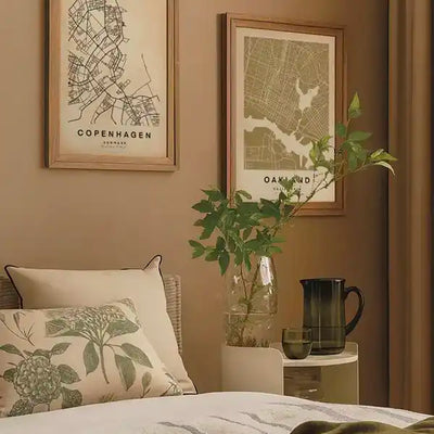 Minimalist bedroom decorated with framed city map posters from the Wanderer Maps™ collection by Cha, featuring Copenhagen and Oakland maps in a soft earthy color palette, paired with natural textiles and greenery.
